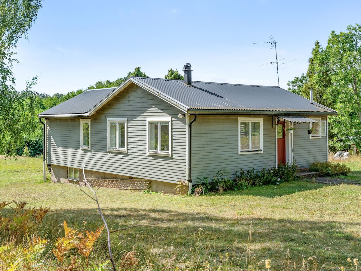 The exterior of the timber house features a combination of grey wood siding and a dark roof. Multiple windows allow natural light to fill the interior. The surrounding grassy area is dotted with trees and includes a pathway leading to the entrance.