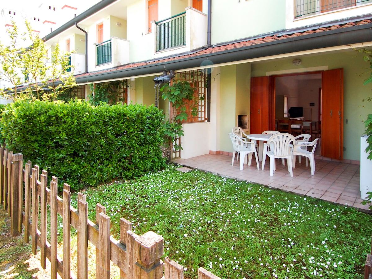 A small garden area features neatly trimmed grass with scattered flowers. Outdoor furniture, including a dining table and plastic chairs, is positioned on a tiled terrace, allowing for relaxation. Green hedges and a wooden fence provide a sense of privacy.
