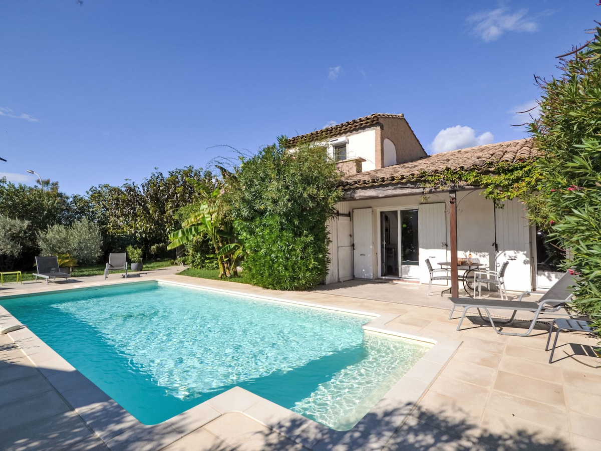 A private swimming pool sits surrounded by lush greenery and a spacious patio area. Loungers and outdoor seating invite relaxation, while the house stands in the background with light-colored walls and tiled roof. Clear blue skies provide a serene backdrop.