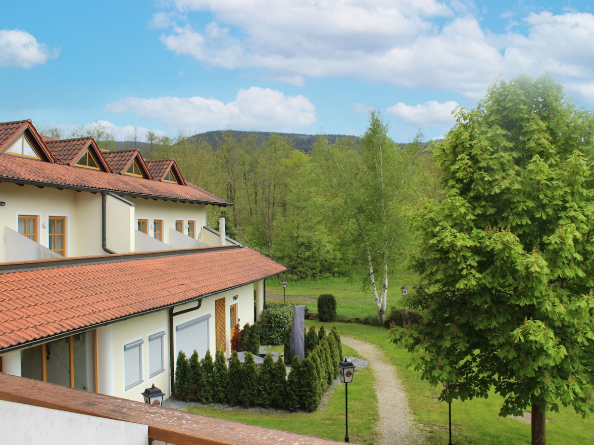 A view of the property reveals a well-maintained building with a red-tiled roof, surrounded by greenery and trees. A gravel path leads through the landscaped area, providing a tranquil setting with mountains in the background and a clear blue sky above.