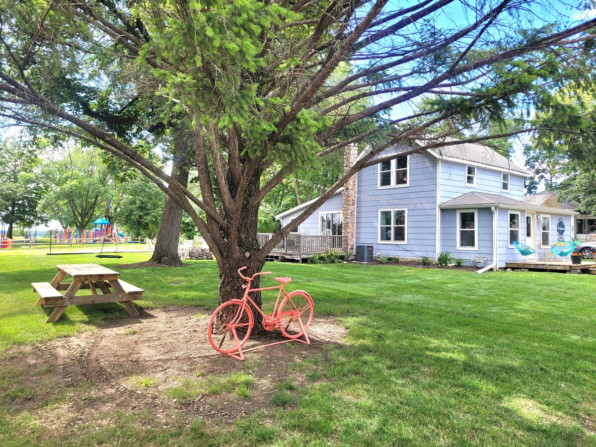 A spacious yard is surrounded by mature trees, featuring a picnic table under a tree and a vintage bicycle painted in coral. The house, with light blue siding and a welcoming porch, is visible in the background, while playground equipment is seen in the distance.