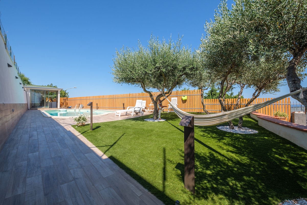 A private outdoor area includes a pool and a green lawn surrounded by olive trees. A hammock is suspended between two posts, and sun loungers are placed nearby. A clear blue sky provides a bright backdrop, enhancing the serene ambiance of the space.