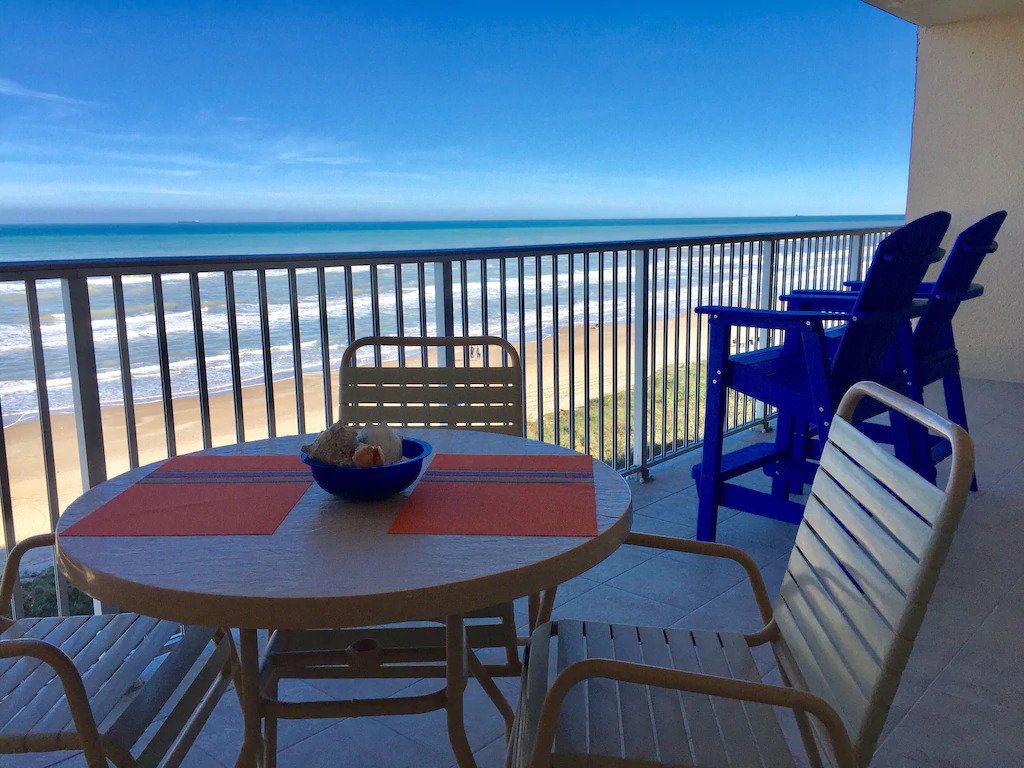 A spacious balcony offers a view of the shoreline with gentle waves lapping against the sand. A round table is set with a bowl, and four chairs are arranged nearby, complemented by high seating overlooking the beach. The ocean and clear blue sky create a serene backdrop.