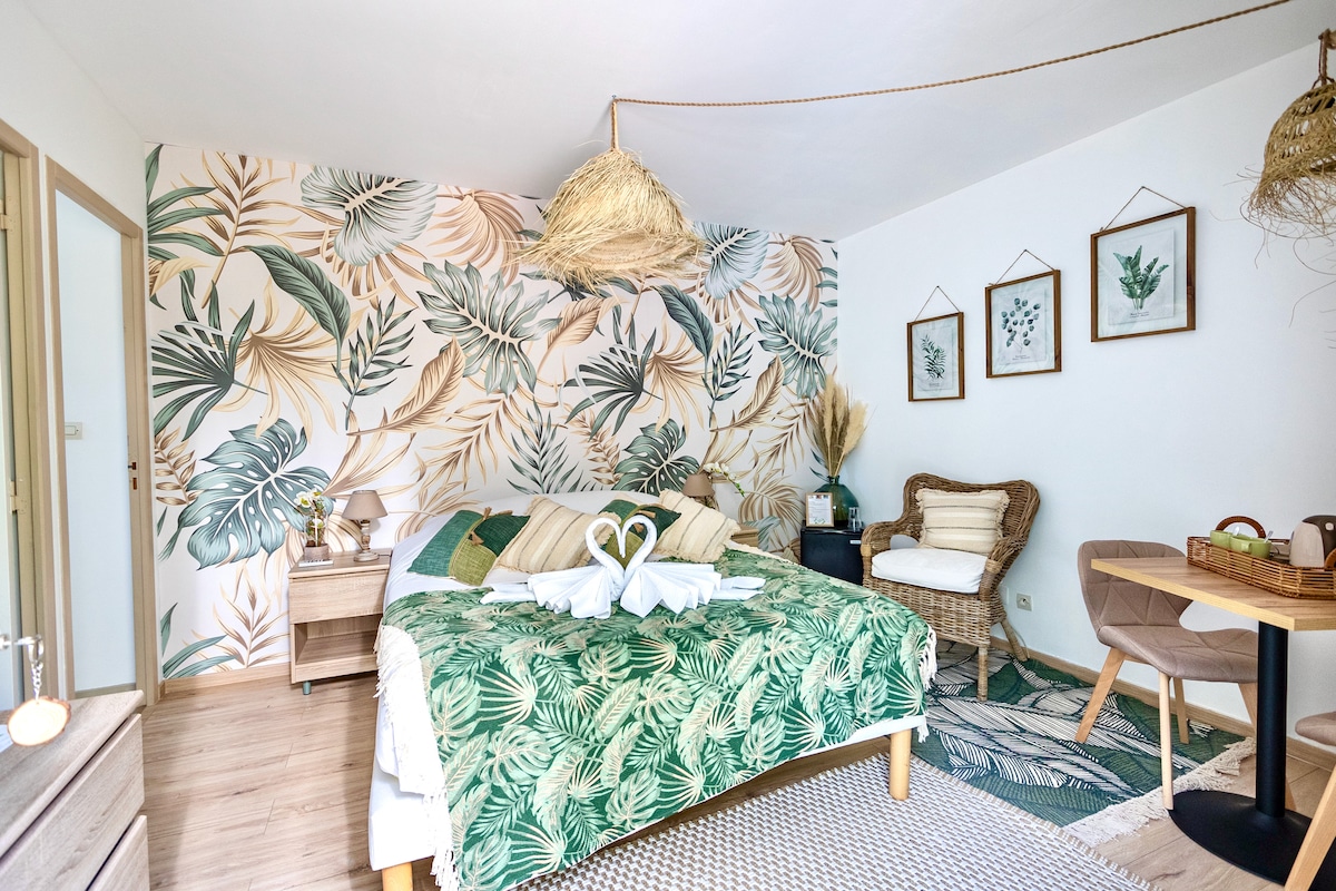A tranquil room features a spacious bed adorned with tropical leaves, complemented by a green decorative throw. Rattan furniture includes a cozy chair and a small table. Botanical wall art and a natural straw light fixture enhance the calming atmosphere.