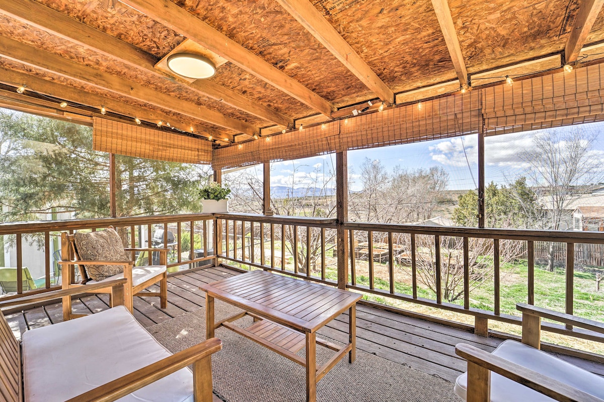A screened patio is highlighted by wooden beams and furniture, including a wooden coffee table and two cushioned chairs. The view beyond features a landscaped yard and trees, with sunlight filtering through the mesh, creating a relaxed outdoor seating area.