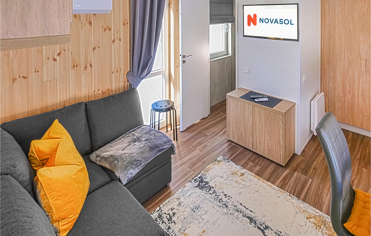 A cozy living area features a gray sofa adorned with colorful cushions and a soft throw. A small side table stands nearby, while a television is mounted on the wall opposite a wood-paneled storage unit. Natural light filters through a window, enhancing the inviting ambiance.