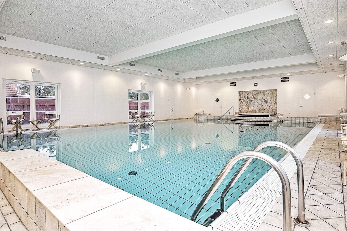 An indoor pool area is presented with a large, clear blue water surface reflecting soft lighting from the ceiling. The pool is bordered by smooth, light-colored tiles. Several lounge chairs are positioned around the poolside, and large windows allow natural light to fill the space.