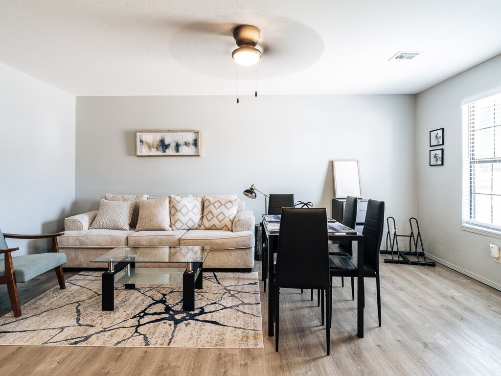 A modern living area features a cozy beige sofa adorned with patterned cushions, complemented by a glass coffee table. Nearby, a dining table with black chairs offers seating for four. Natural light streams in through a window, highlighting the textured area rug and minimalist décor.