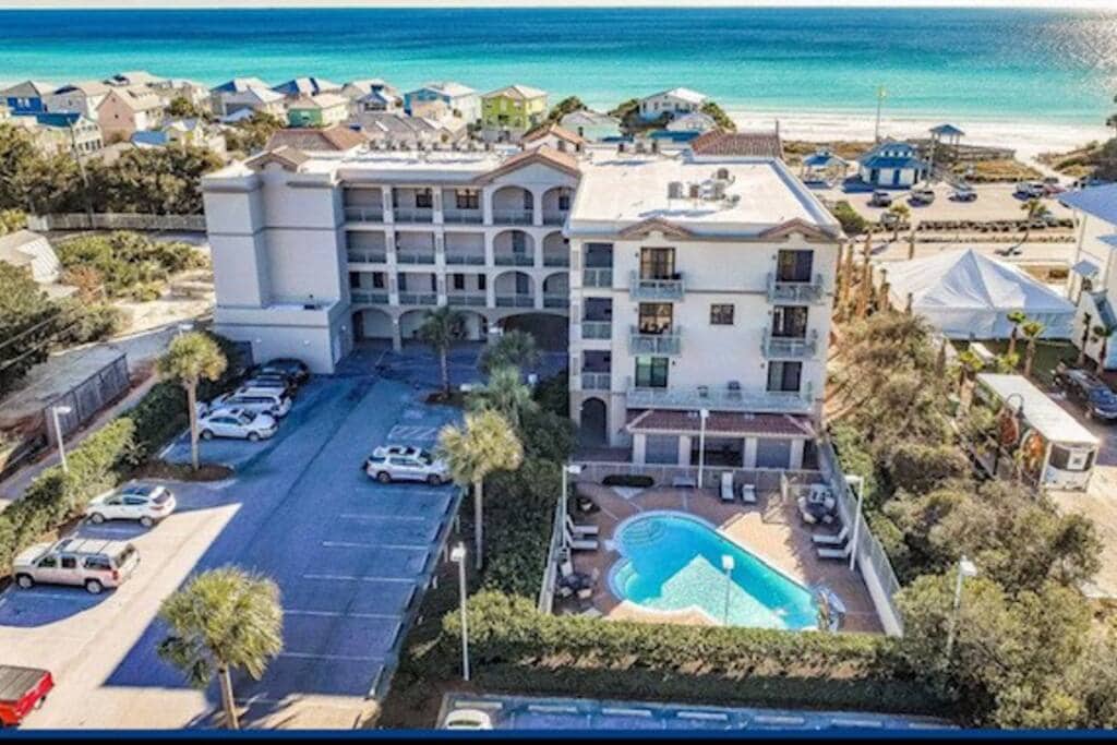 An aerial view of a coastal condominium property is displayed, showcasing a refreshing pool surrounded by palm trees. The Gulf of Mexico is visible in the background, along with nearby buildings and parking spaces. The tranquil setting highlights the property's close proximity to the beach.