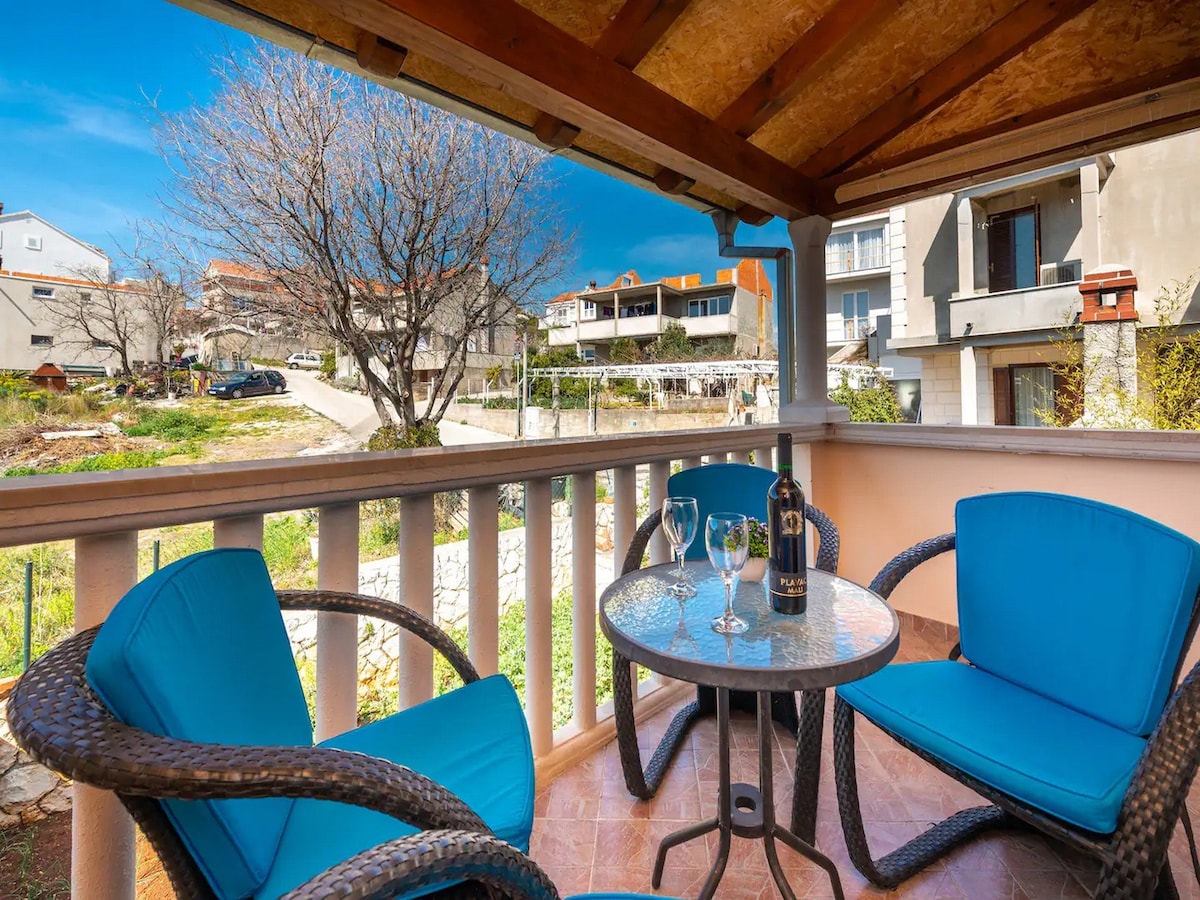 A private terrace is furnished with a circular table and four blue chairs, creating a space for outdoor relaxation. The surrounding area features greenery and buildings, with a clear sky above, enhancing the tranquil atmosphere.