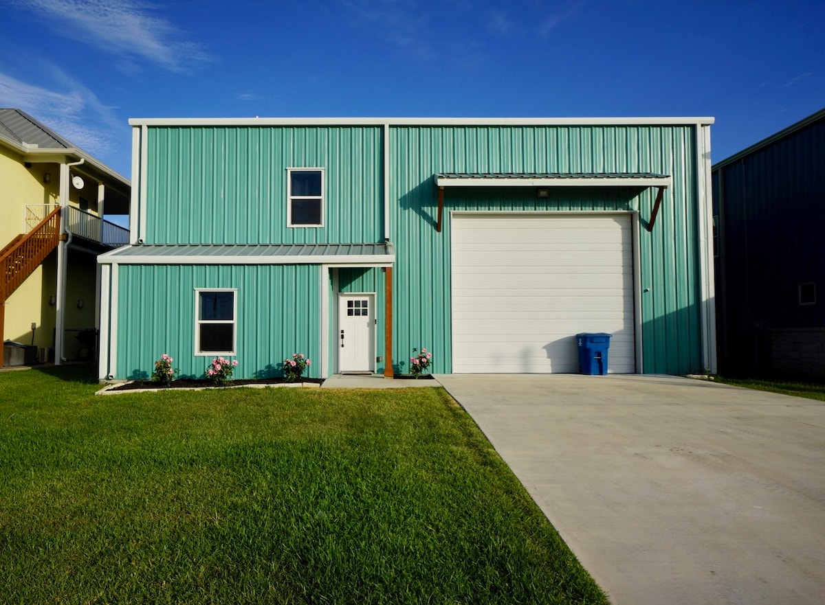 A two-story building featuring a teal metal exterior and a large white garage door. The ground is landscaped with green grass and flower beds near the entrance, contributing to a well-maintained exterior.
