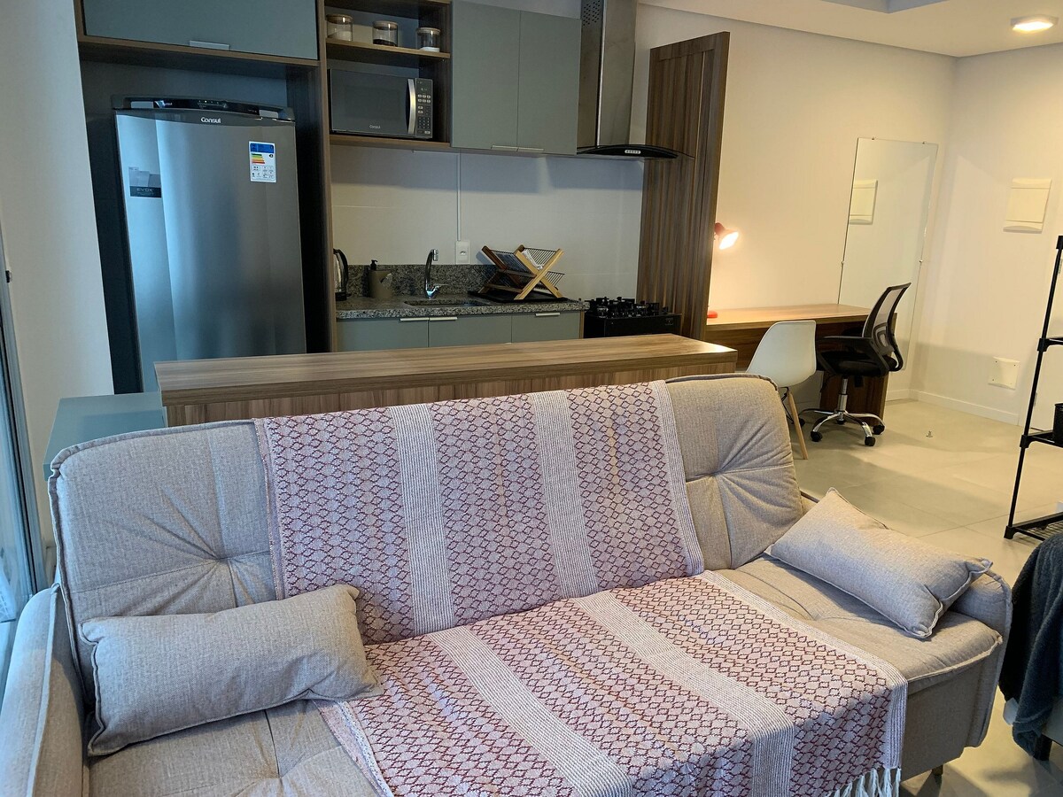 A modern living area features a light-colored sofa adorned with decorative cushions. A compact kitchen is visible in the background, equipped with stainless steel appliances and cabinets. A workspace with a desk and chair is also present, ensuring a functional setup for productivity.