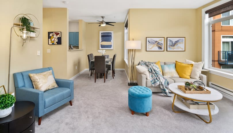 A spacious living area features a light-colored carpet and neutral walls. A comfortable sofa with decorative pillows faces a stylish coffee table. A reading chair is positioned nearby, alongside a bright blue ottoman. The open layout leads to a dining area with a table and chairs.