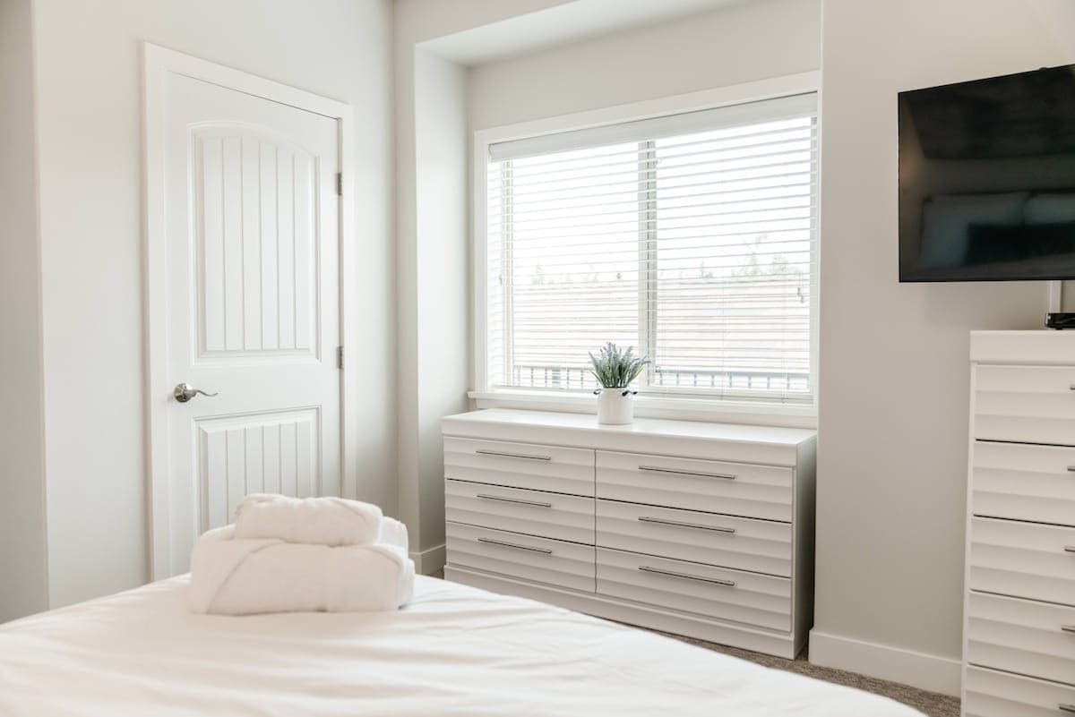A bright and airy bedroom is presented, featuring a spacious bed with neatly arranged towels at the foot. A modern dresser occupies one side, while a window with white blinds allows natural light to fill the room, complemented by a small potted plant on the dresser.