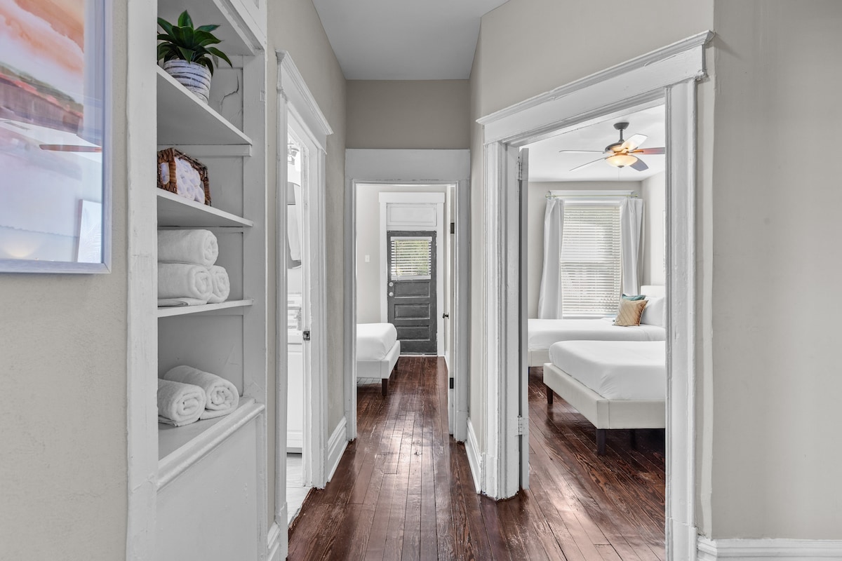 A hallway connects multiple rooms, featuring polished hardwood floors and neutral-toned walls. Shelves lined with neatly folded towels are visible, while doorways lead into well-lit bedrooms. A ceiling fan adds to the comfort, with natural light streaming in through nearby windows.