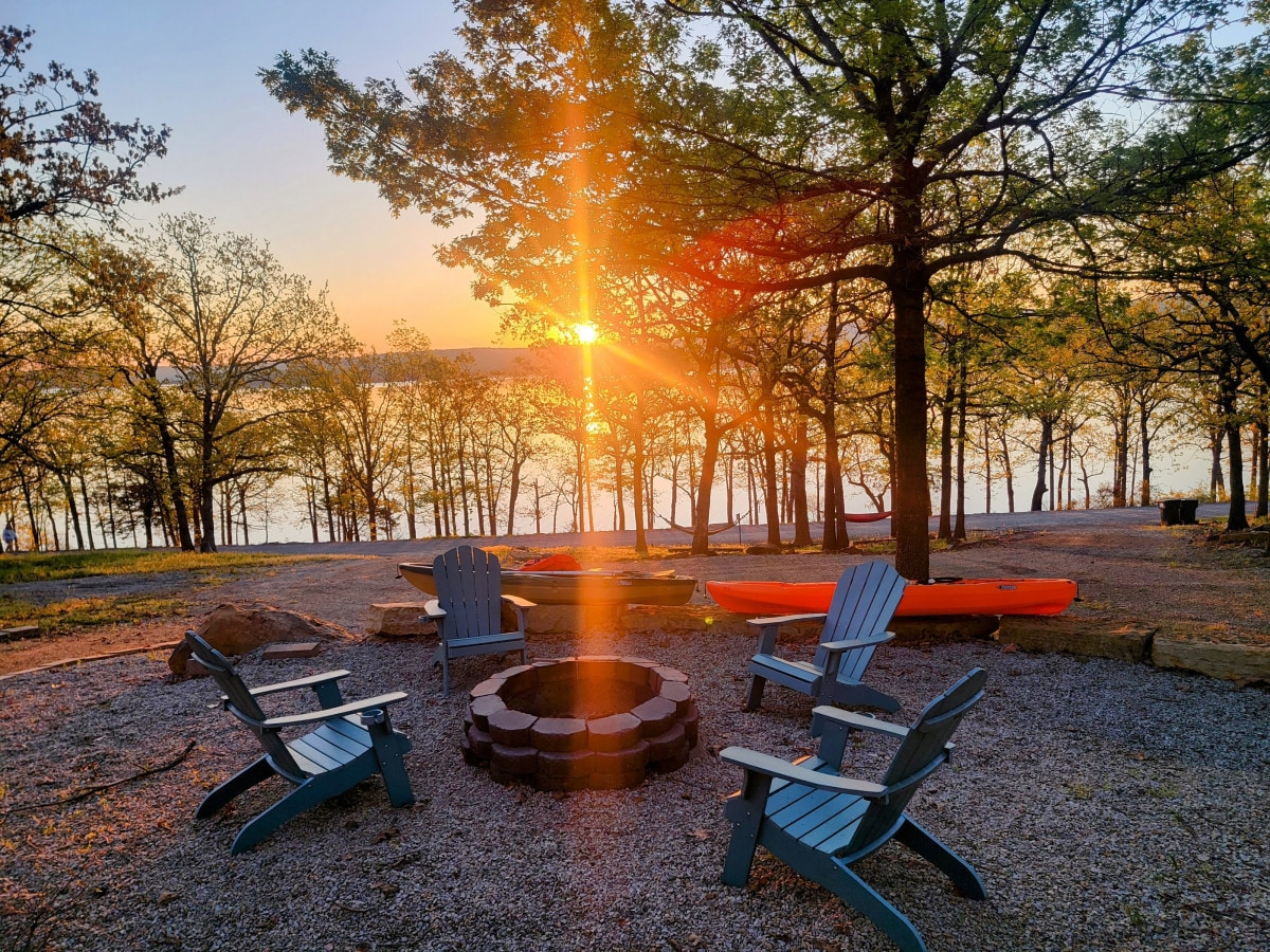 A cozy fire pit area is surrounded by several blue Adirondack chairs, positioned on a gravel surface. Trees frame the scene as the sun sets, casting warm rays over the tranquil lake visible in the background. An orange kayak rests nearby, ready for use.
