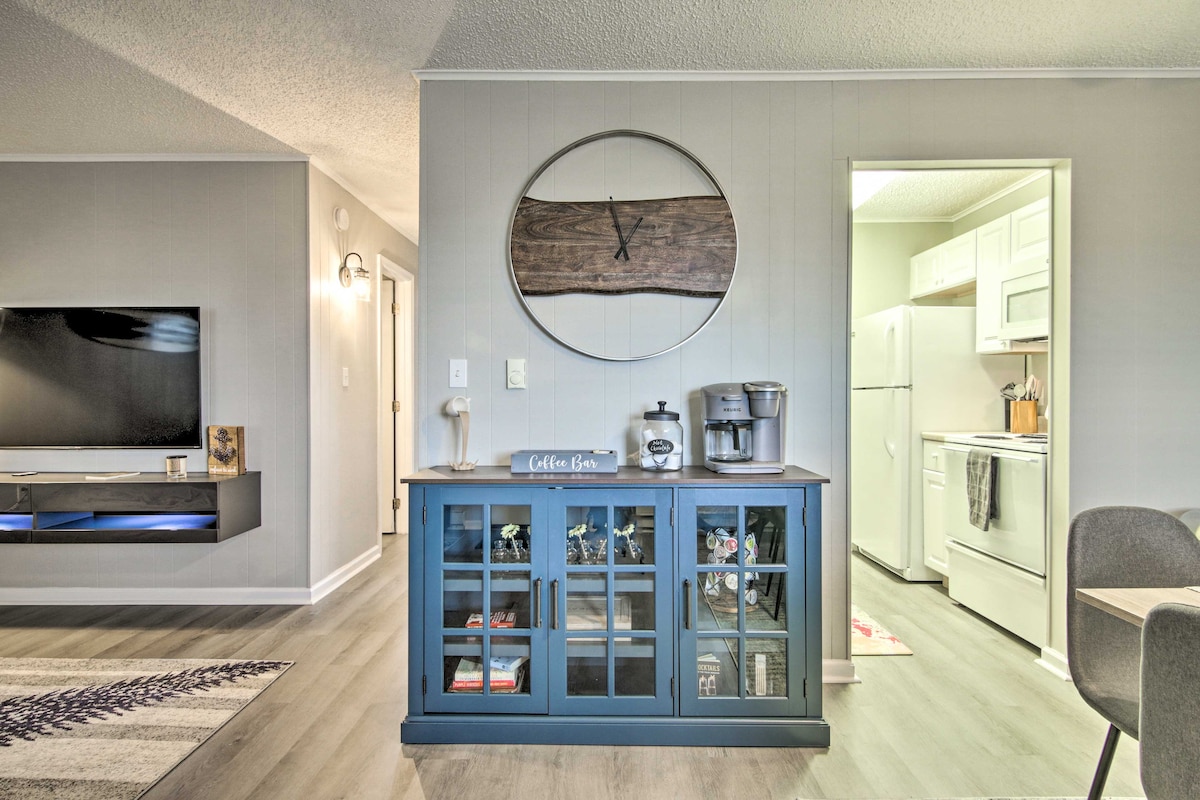 A living area is depicted, featuring a blue cabinet displaying decorative items. A round wall art piece adorned with crossed objects is mounted above the cabinet. An open kitchen area is visible in the background, showcasing white cabinets and modern appliances.