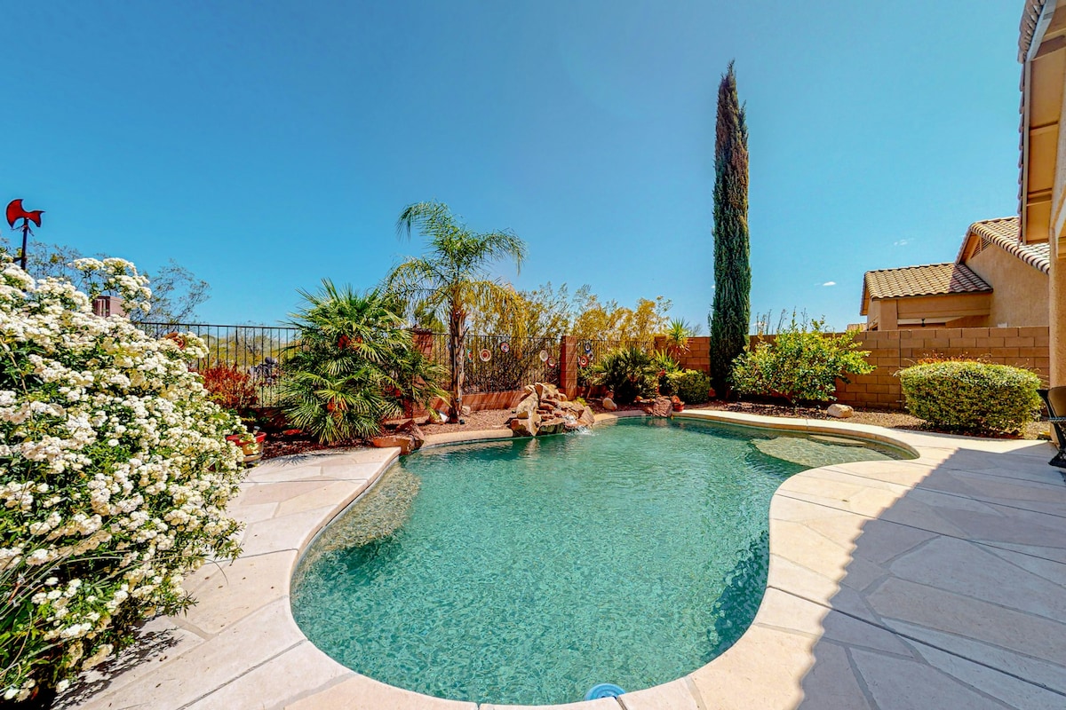 A private pool is surrounded by lush landscaping, featuring various plants and vibrant flowers. The gently curved pool has clear water reflecting the blue sky. A tall cypress tree stands in the background, adding height to the garden view.