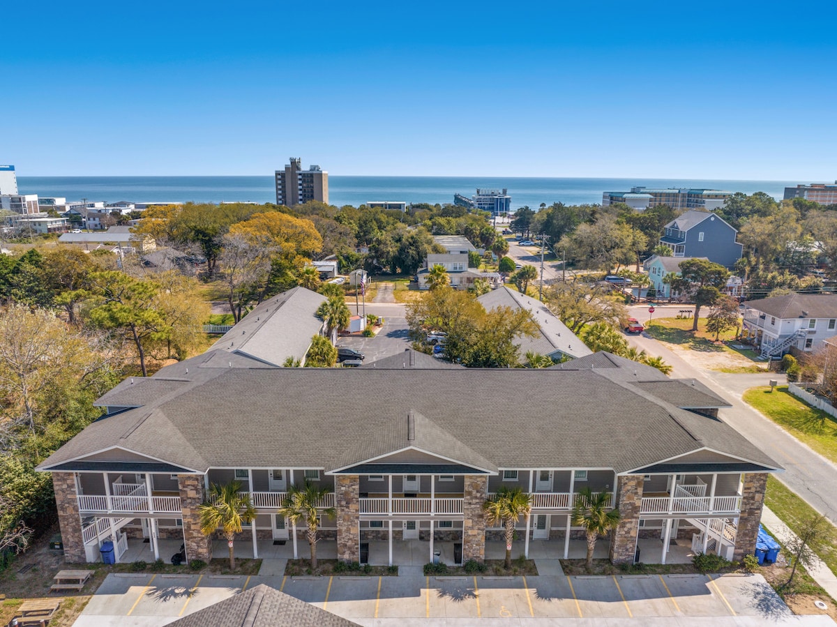 The exterior of a modern apartment complex is depicted, featuring a spacious layout with multiple entrances. Lush greenery surrounds the building, with a clear view of the ocean in the background. The bright blue sky enhances the inviting atmosphere of the area.