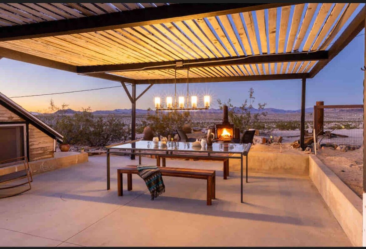 An outdoor dining area features a long glass table surrounded by chairs, under a wooden pergola. A modern chandelier hangs above, illuminating the space, while a wood-burning stove provides warmth. The backdrop showcases desert landscapes under a twilight sky.
