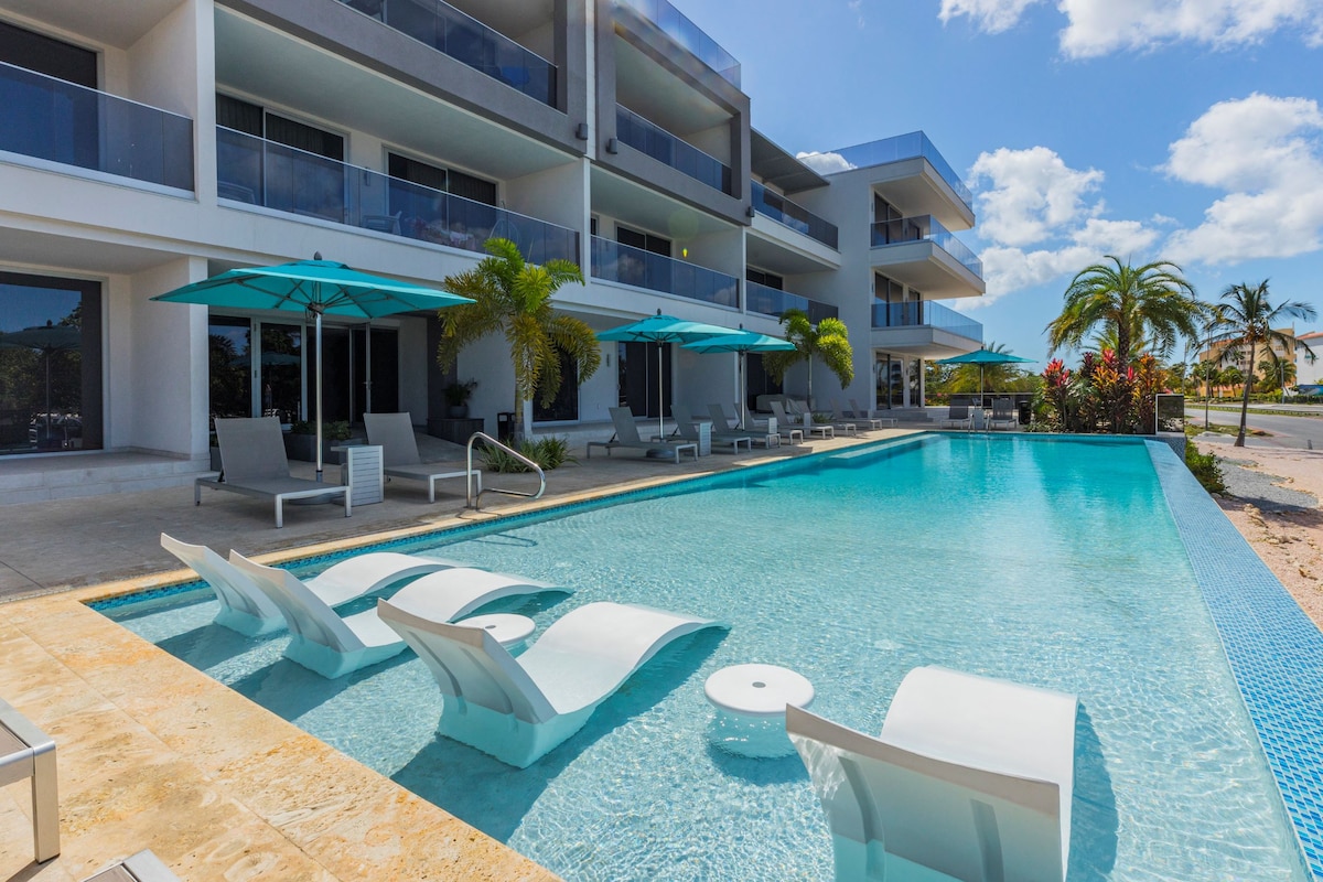 An infinity pool with built-in sunbeds invites relaxation, bordered by a spacious terrace. Sun umbrellas provide shade, while palm trees and blooming plants enhance the outdoor ambiance. The modern condominium structure features large glass doors, allowing for unobstructed views of the pool and surrounding area.