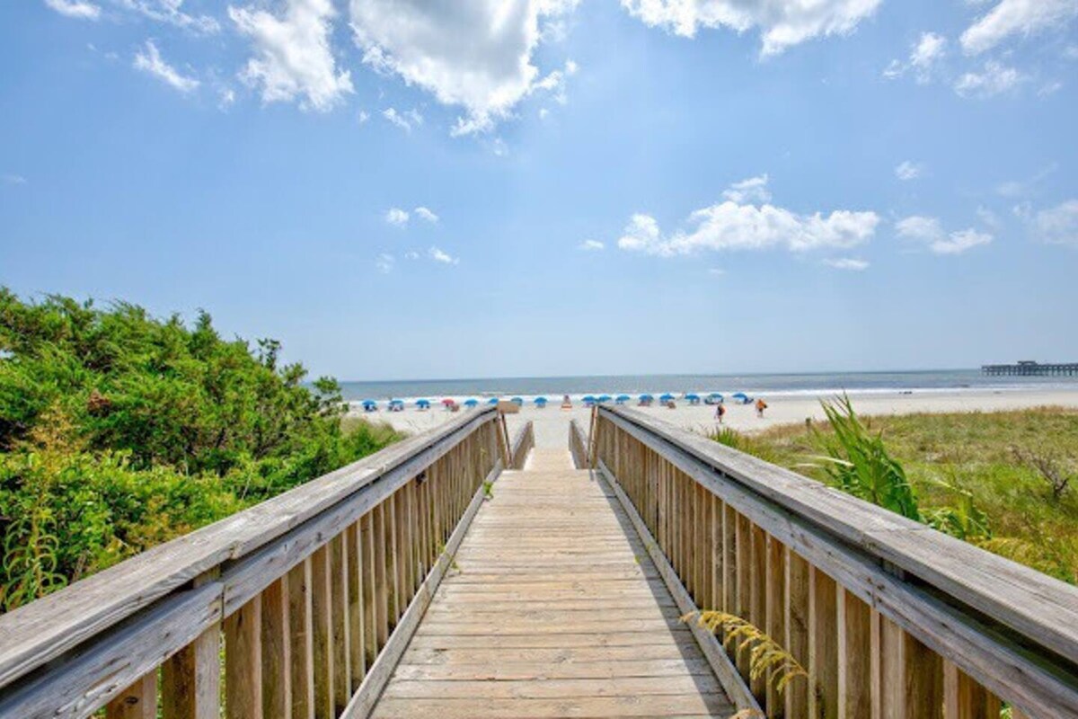 A wooden boardwalk leads directly to the sandy beach, flanked by green foliage on either side. The distant ocean is visible, dotted with colorful beach umbrellas. A clear blue sky, punctuated by a few clouds, creates a bright and inviting scene.