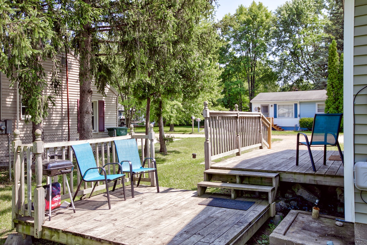 An outdoor deck is visible, featuring two blue chairs positioned for relaxation. Surrounding greenery provides a natural backdrop, while the nearby steps lead to a welcoming entryway. The area conveys a peaceful ambiance, ideal for enjoying the outdoors.
