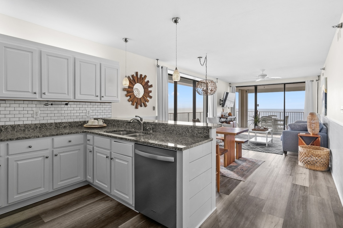 The spacious kitchen features light grey cabinetry, a granite countertop, and modern appliances. Natural light floods the open area, revealing a dining space with a table, while views of the beach are visible through large sliding glass doors. The adjacent living area includes comfortable seating.