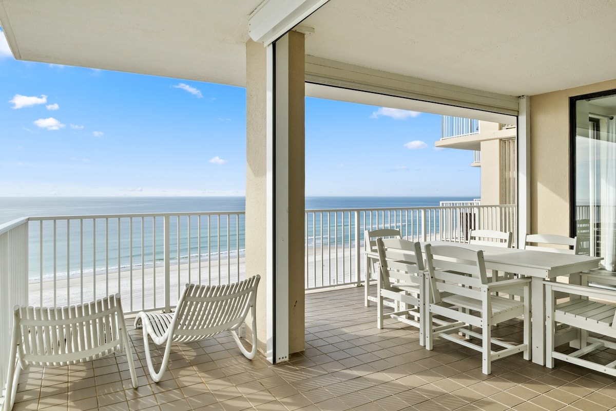 A spacious private balcony offers panoramic views of the Gulf of Mexico. Outdoor seating includes a large dining table and lounge chairs, ideal for enjoying the coastal breeze. The floor-to-ceiling sliding doors provide easy access to the stunning beachfront scenery.