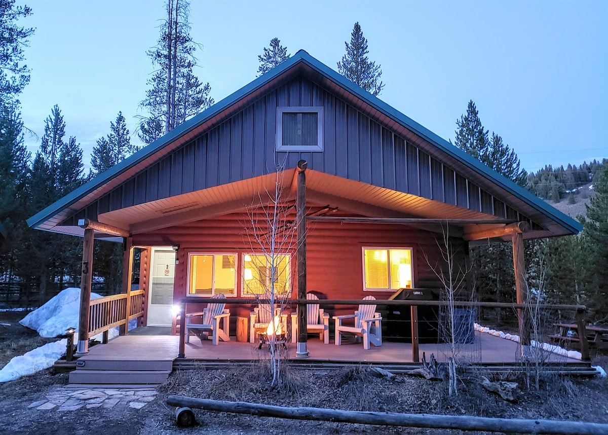 The cabin exterior is shown at dusk, revealing warm light glowing from the windows. A covered patio features several wooden lounge chairs and is framed by surrounding trees. A pathway leads to the entrance, enhancing its inviting presence in the natural setting.