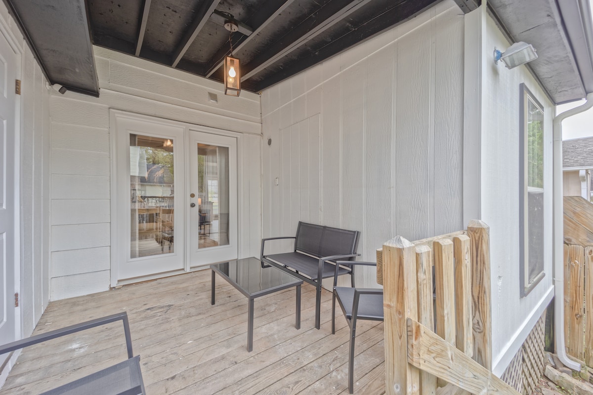 An outdoor seating area is presented with a glass-top table and two metal-framed chairs. The wooden deck is partially covered, and large glass doors offer a view into the interior space. A pendant light hangs from the ceiling, providing illumination.
