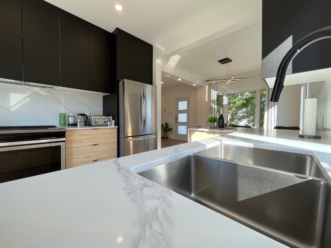 A modern kitchen is depicted, featuring dark cabinetry with clean lines, a stainless steel refrigerator, and a large marble countertop. A double sink is visible, along with a sleek faucet. Natural light enters through large windows, illuminating the space.
