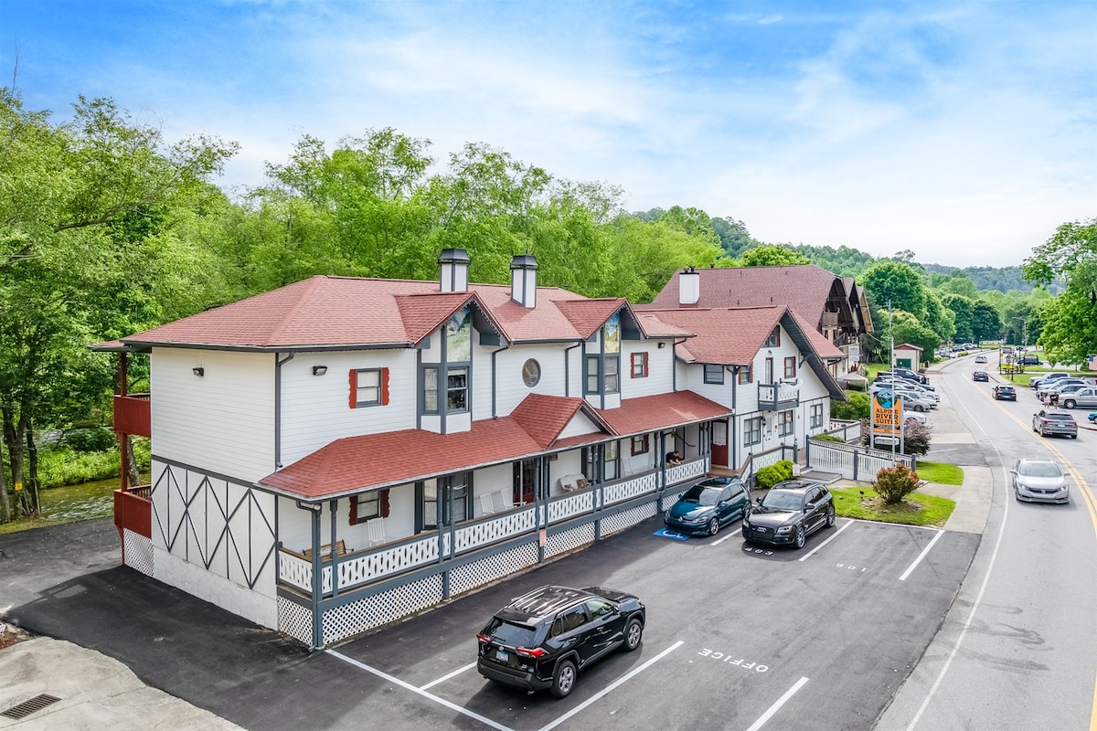 The exterior of a three-story building is visible, featuring a combination of white walls and red accents. A pitched roof and decorative eaves complement the structure. The parking lot accommodates several vehicles, and lush greenery is seen in the background, with a glimpse of the road in front.