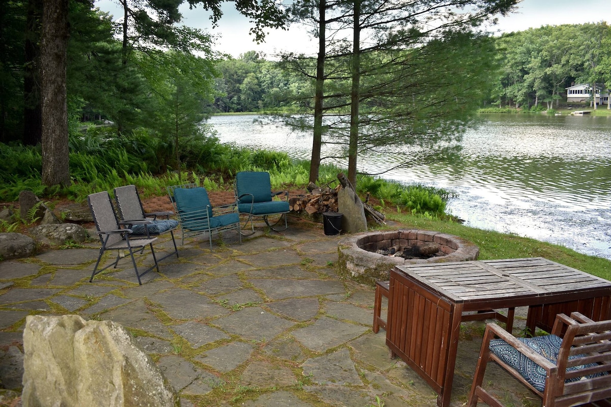 A flagstone patio along the water features a fire pit surrounded by folding chairs. A wooden table is positioned nearby, with a serene lake view and lush greenery in the background. The space offers a tranquil setting for relaxation and outdoor dining.