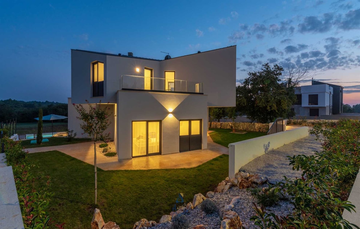 The modern villa is illuminated against a twilight sky, highlighting its unique architectural design. Soft light spills from the windows, reflecting off the smooth surfaces. The surrounding garden features manicured grass and gravel pathways, leading to a private pool visible in the background.