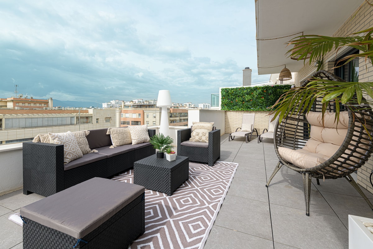 An outdoor terrace features modern garden furniture, including a sectional sofa and a hanging chair. A geometric patterned rug is placed beneath a lighted lamp, and potted greenery adds a touch of nature. City views are visible against a cloudy sky.