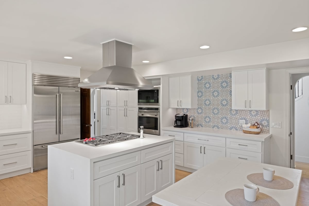A bright kitchen with modern cabinetry features a large central island equipped with a gas cooktop. Stainless steel appliances are integrated, while a decorative blue and white tiled backsplash adds a subtle design element. Ample natural light is provided through overhead recessed lighting.