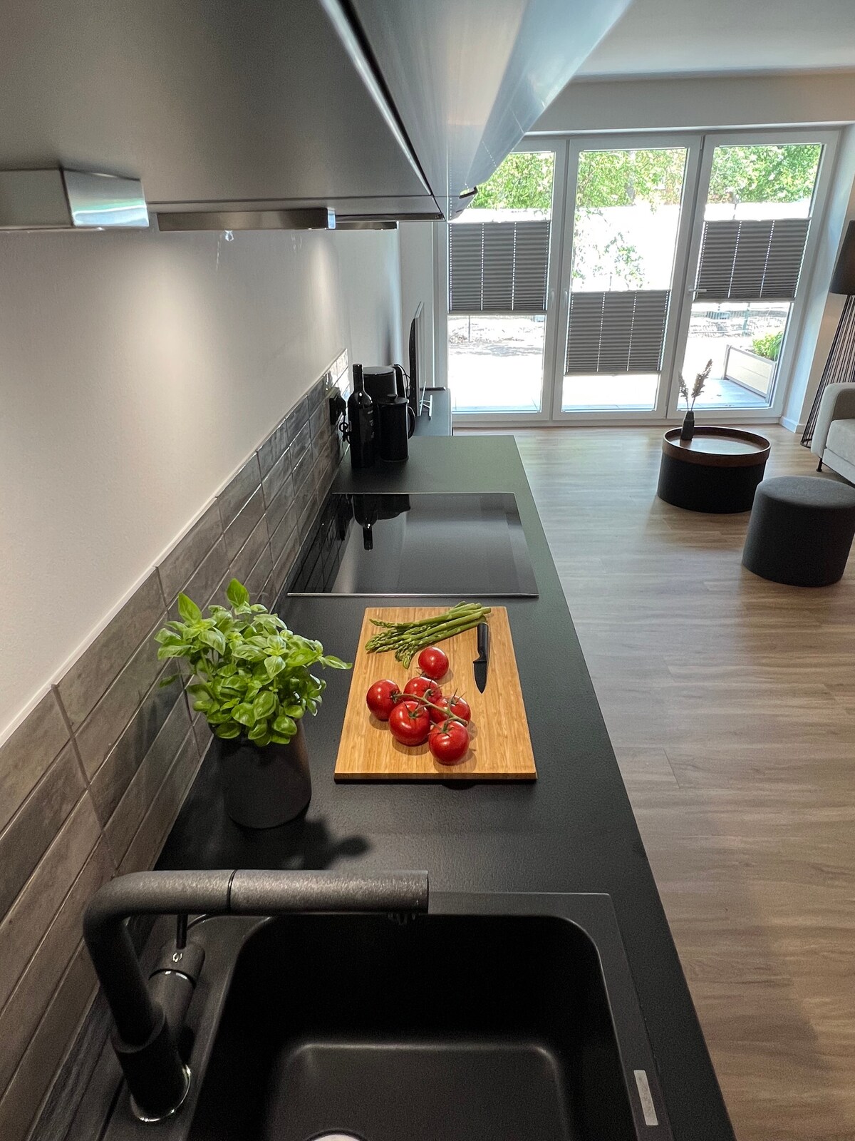 A modern kitchenette features sleek black countertops and cabinetry, complemented by a wooden cutting board with fresh tomatoes and herbs. Large windows provide natural light, with shutters partially closed, creating a bright and functional cooking space.