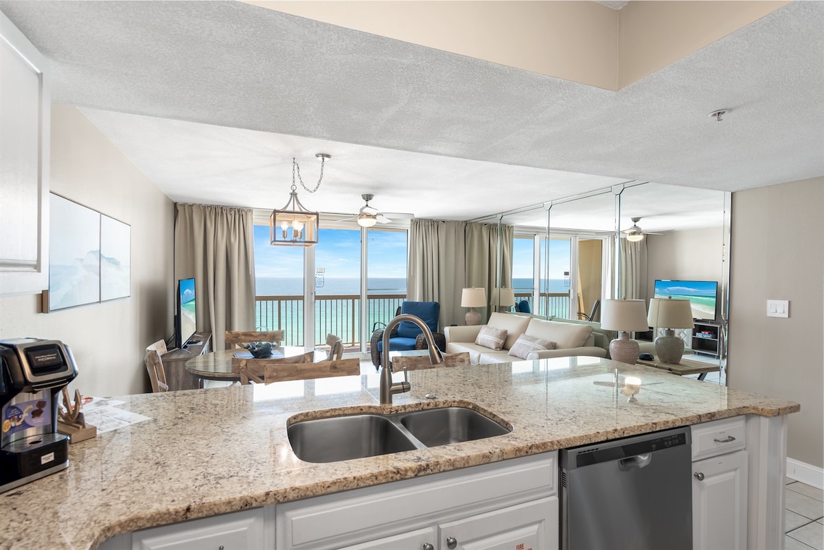 The open-concept living area features a kitchen with granite countertops and stainless steel appliances, including a dishwasher. The space is filled with natural light from large glass doors leading to a balcony with ocean views, offering comfortable seating and a welcoming atmosphere.