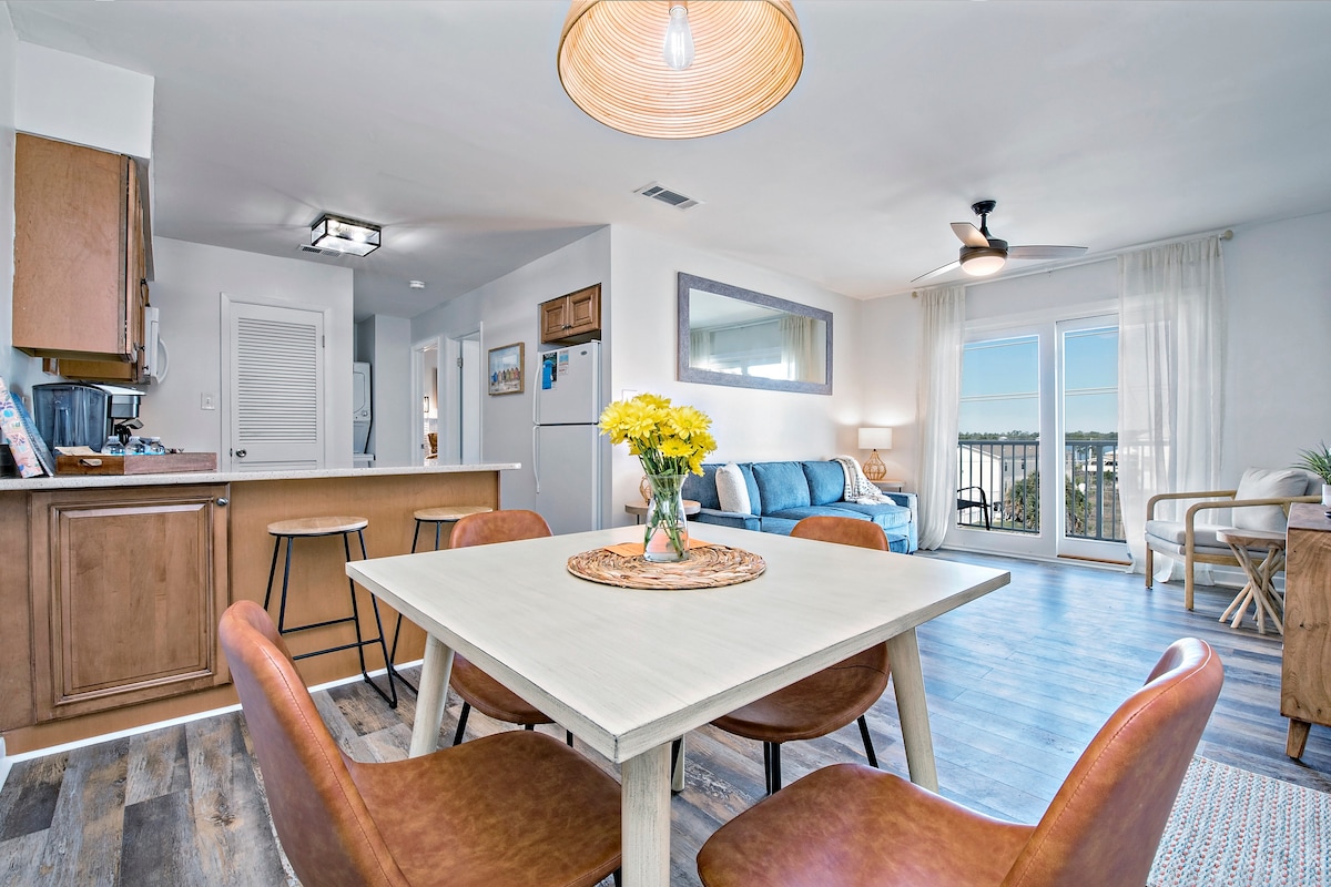 A dining area features a light wooden table surrounded by four brown chairs. A vase of yellow flowers sits at the center. The space opens to a bright living area with a blue sofa and large glass doors leading to a balcony, offering views of the lagoon.