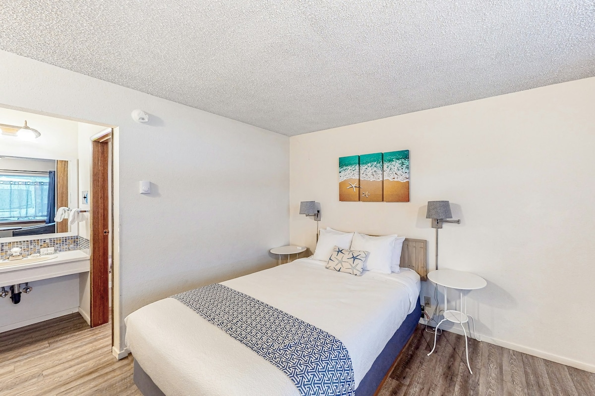 A bright guest room features a spacious bed with a white duvet and decorative blue throw. Two bedside tables with lamps are positioned beside the bed. A sea-themed artwork adorns the wall, and a bathroom area is visible in the background.