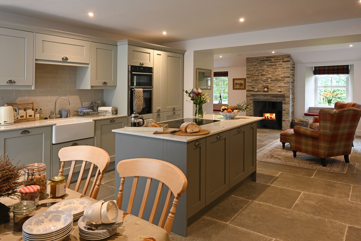 An open-plan kitchen is equipped with modern appliances and grey cabinetry. A large island provides additional workspace alongside a farmhouse dining table set for guests. A cozy living area is highlighted by a stone fireplace, with comfortable seating that encourages relaxation.