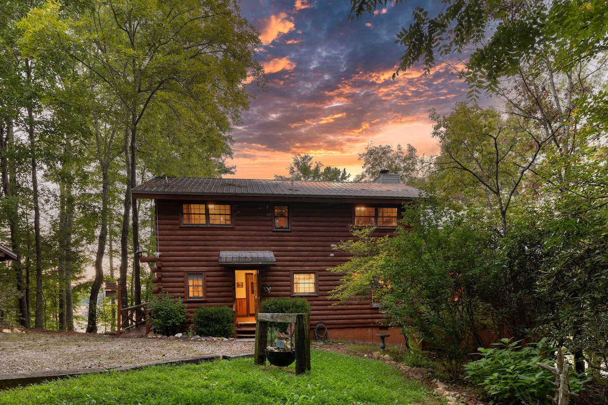 The exterior of a two-story log cabin is depicted, nestled among trees with a gravel pathway leading to the entrance. Warm light glows from several windows, while the vibrant sunset casts hues across the sky. A hint of the lake is visible beyond the cabin.
