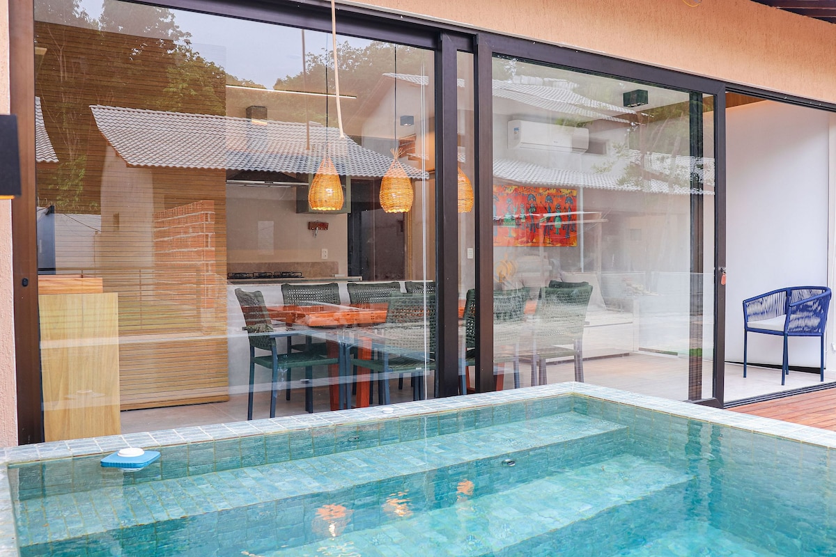 A private pool is showcased in the foreground, bordered by smooth stone. Expansive glass doors offer a view into the interior dining area, featuring a table and chairs. Warm light is emitted from pendant fixtures above, highlighting a cozy atmosphere.