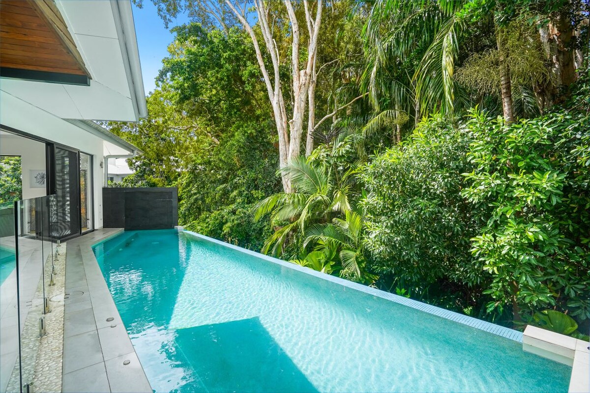 An inviting wet edge pool extends alongside the house, surrounded by lush greenery. The tranquil water reflects the sunlight, creating a serene atmosphere. Tall trees and dense foliage frame the scene, enhancing the sense of privacy and connection to nature.