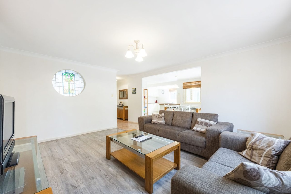 A bright living area is featured, showcasing two cushioned sofas arranged around a wooden coffee table. Soft lighting is provided by three pendant lights above, while a decorative window allows natural light to enhance the space. A view of the adjoining kitchen can be seen in the background.