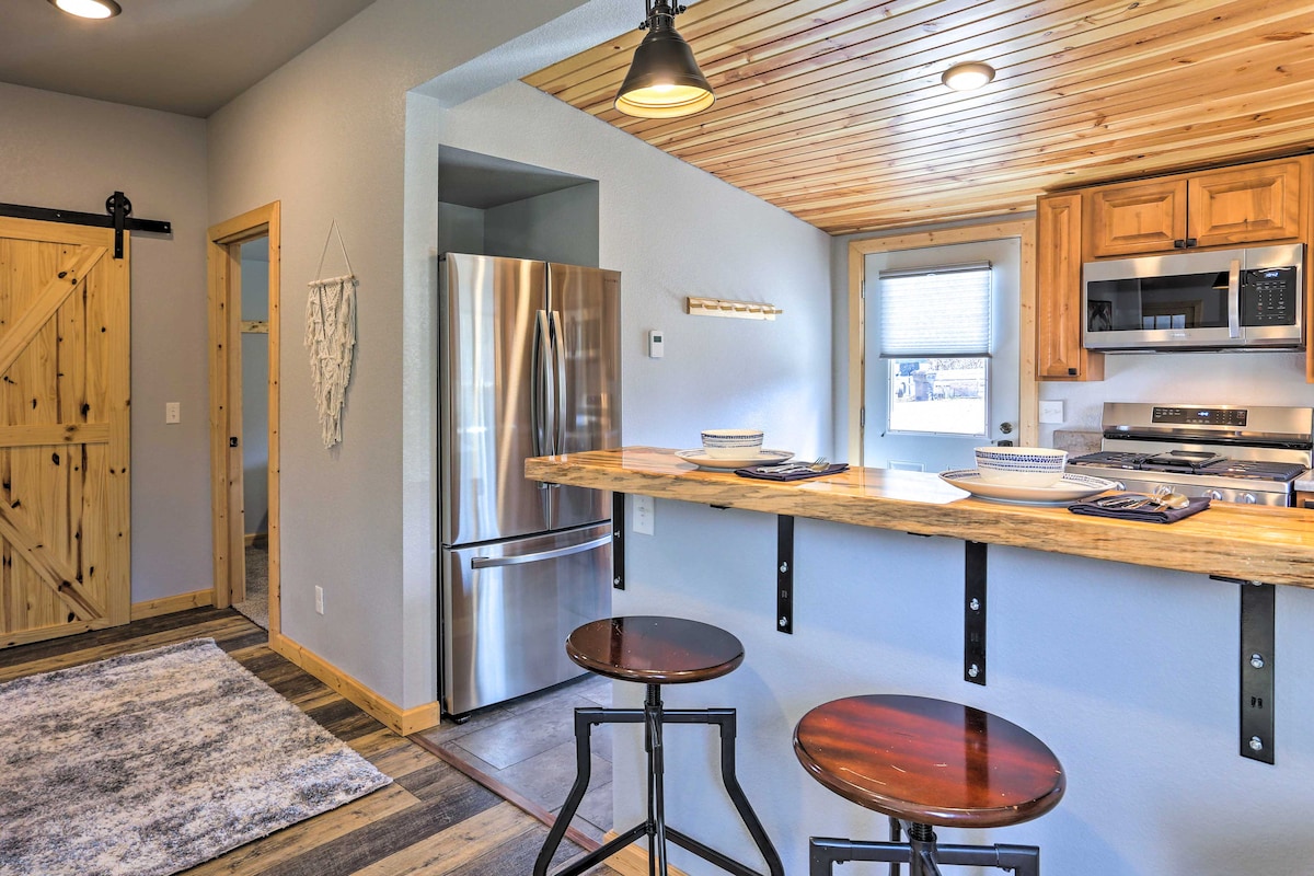 A modern kitchen space is highlighted by a wooden countertop that extends into a bar area with two metal stools. Stainless steel appliances, including a refrigerator and microwave, are visible. Light wooden cabinetry complements the natural wood ceiling, enhancing the clean and functional design.