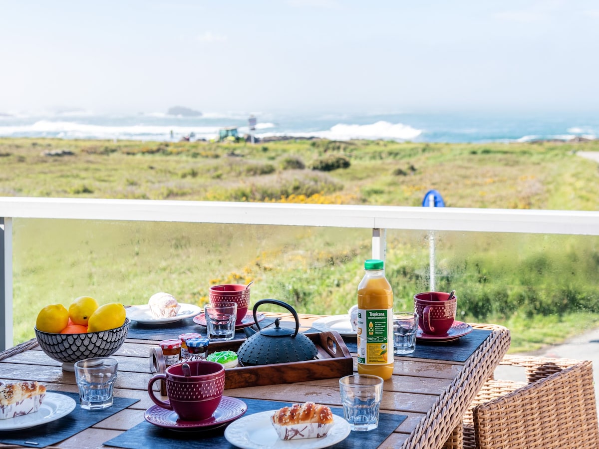 A balcony table is set for breakfast, featuring colorful dishes, glasses, and a bottle of juice. Lush greenery and a view of the sea create a serene backdrop, while distant waves and a coastal landscape add to the inviting atmosphere.