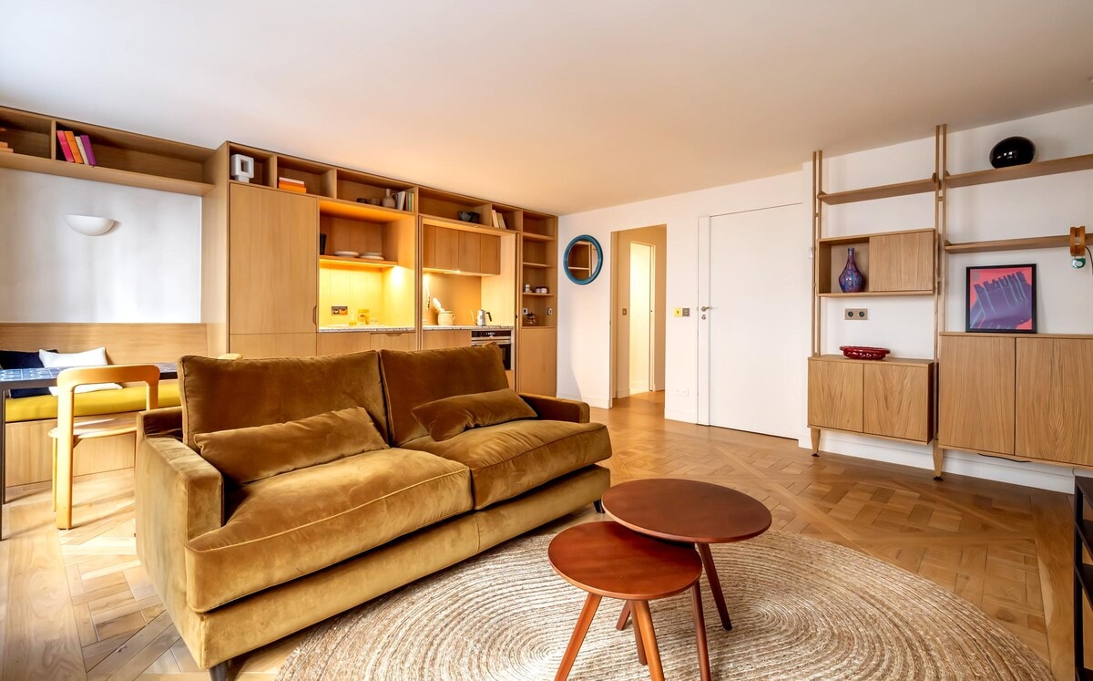 A spacious living area features a plush, earth-toned sofa paired with two round wooden tables on a textured area rug. Shelving units along the walls display decorative items and books, while soft lighting adds warmth to the neutral decor.