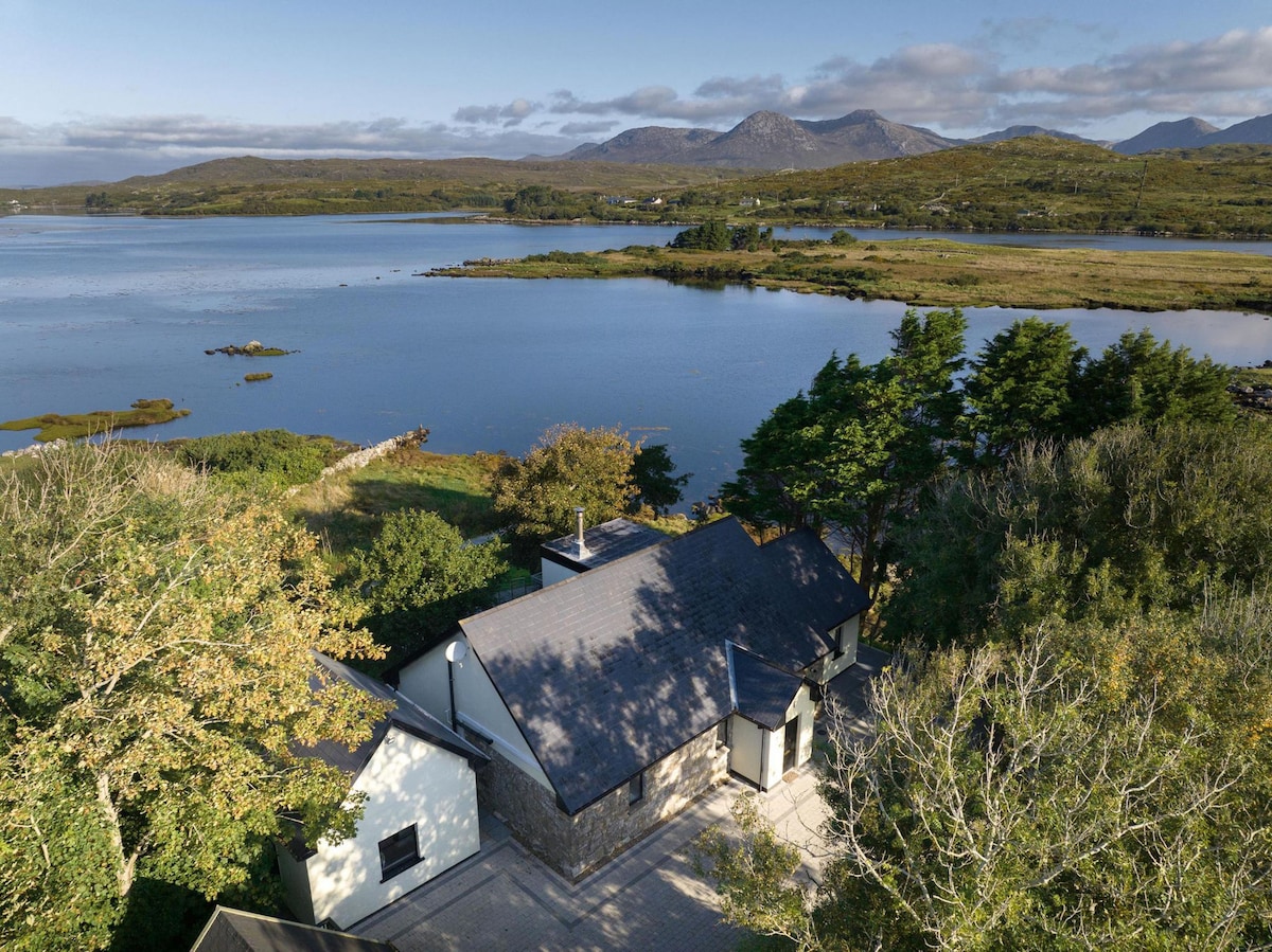 An aerial view of the cottage showcases its modern design nestled among lush greenery, with a backdrop of serene waters and distant mountains. The surrounding landscape offers a peaceful ambience, reflecting the natural beauty of Connemara.